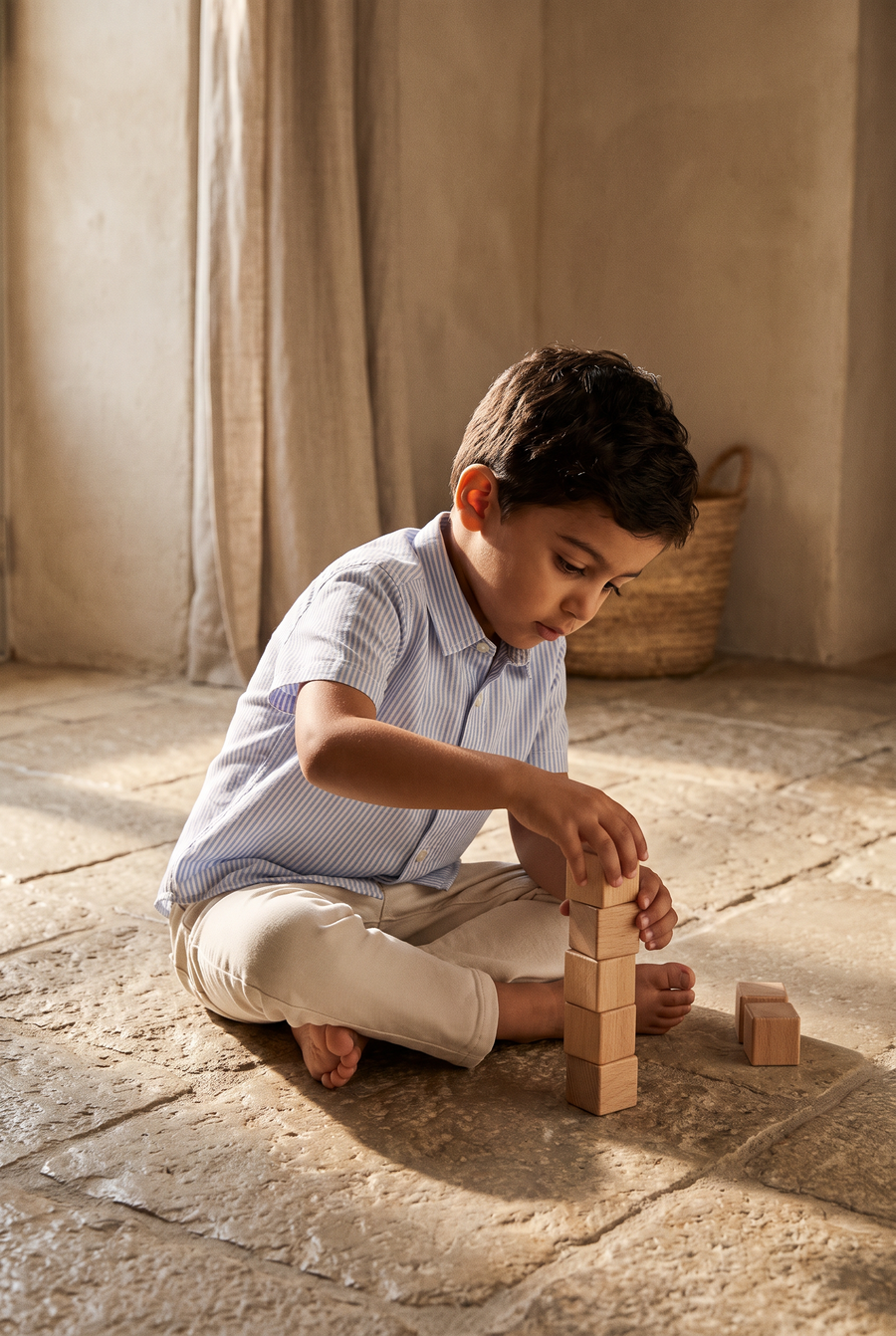 a boy sitting on a stone floor stacking wooden blocks, in the coastal blue shirt and salty white trousers