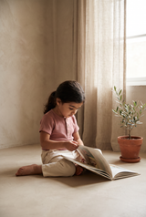 a girl sitting on the floor turning the page of a picture book, in the pink pepper tee and salty white trousers